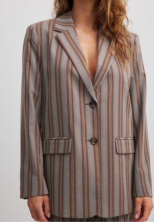 Kvinde iført en brun og grå blazer med lodrette striber, to knapper og lommer med klap, stående foran en ensfarvet baggrund.