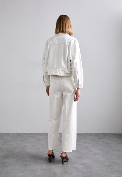 Weißer, kurzer Jeansjacke im Oversize-Schnitt, kombiniert mit weißen weiten Jeans mit ausgefransten Säumen. Braune Absätze runden den Look ab.