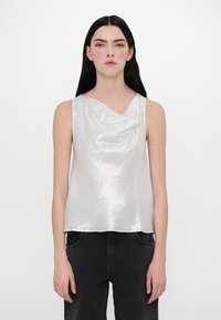 Top sans manches en tissu argenté brillant, avec un col drapé et une coupe décontractée. Porté avec un pantalon en denim foncé, au design simple et à la texture lisse.