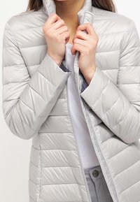 Kobieta w jasnoszarej, pikowanej kurtce puchowej, trzymająca kołnierz obiema rękami, częściowo zapięta nad białą koszulą.