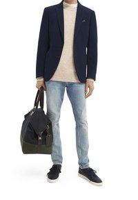 Blazer azul marino, suéter de punto beige, jeans de luz azul y zapatillas oscuras. Sosteniendo un bolso de lona oscura y una bolsa de deporte verde con detalles en cuero marrón.