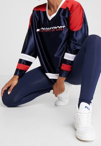 Camiseta deportiva azul marino con detalles de malla en rojo y blanco, con cuello en V y logo. Combinada con leggings azul marino y zapatillas blancas.
