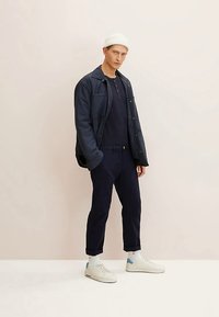 Abrigo azul marino abotonado sobre un suéter azul marino, combinado con pantalones azul marino y zapatillas blancas con detalles azules. Modelo lleva un gorro blanco.