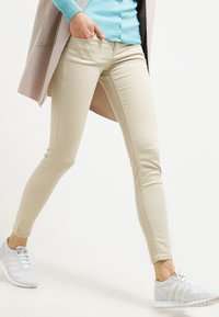 Jeans skinny beige hechos de tejido elástico, con una textura suave y un diseño de ajuste ceñido, combinados con zapatillas deportivas grises.