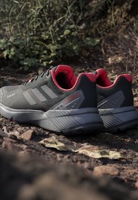 Scarpe sportive nere con accenti rossi, tomaia in rete testurizzata, tallone rinforzato e suola in gomma grigia, progettate per l'uso all'aperto.