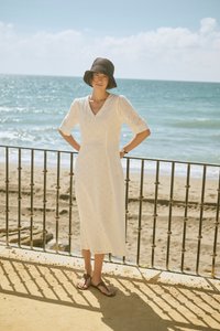 Žena u bijeloj haljini s perforacijama i crnom šeširu stoji kraj metalne ograde s oceanom i pješčanom plažom u pozadini na sunčan dan.
