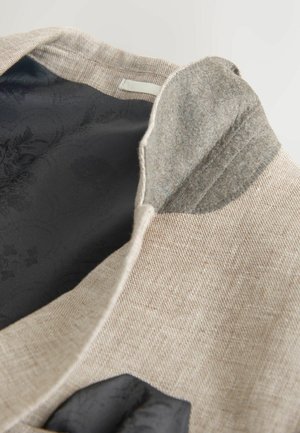 Světlý béžový lněný kabát se šedým plstěným límcem a černou vzorovanou vnitřní podšívkou. Součástí je malý kapesník z tmavé látky.