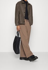 Veste marron texturée avec un col, chemise noire, pantalon large beige et chaussures noires. Sac fourre-tout noir avec poignées.