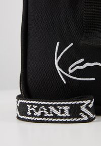 Borsa in tessuto nera con logo ricamato bianco. Presenta una superficie texturizzata e una tracolla distintiva con scritte bianche e cuciture decorative.