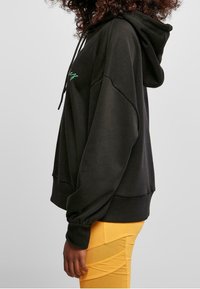 Černá mikina s kapucí a stahovací šňůrkou, se zeleným vyšívaným textem. Má široké rukávy a volný střih, doplněná o žluté kalhoty.