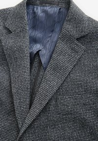 Struktureret grå blazer med sorte detaljer, der har en klippet krave og en silkeblåt mønstret foring. Nærbillede af indersiden.