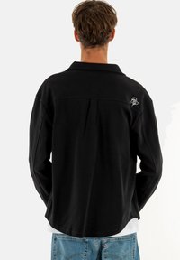 Chemise à manches longues noire avec un design col, comportant une couture dans le dos et un logo brodé sur l'épaule.