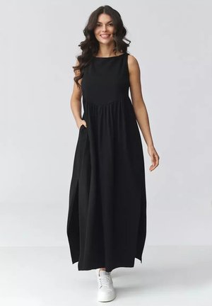 Uśmiechnięta kobieta z długimi kręconymi włosami, ubrana w czarną, bezrękawną sukienkę maxi i białe trampki, stojąca na jednolitym tle.