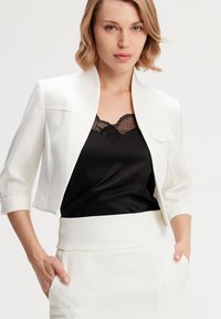 Veste blanche courte avec un col rond, un design structuré et des manches retroussées, associée à un haut en satin noir avec des accents en dentelle.