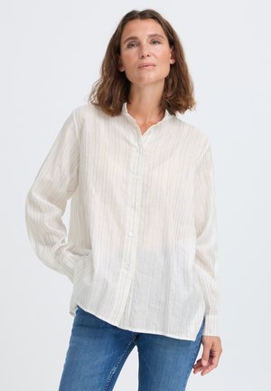 Woman with shoulder-length brown hair wearing a loose white striped button-up shirt and blue jeans standing with one hand in pocket.