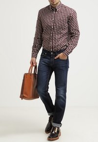 Man med mönstrad långärmad skjorta, mörka jeans med uppvikta ärmar, mörka läderskor, som håller en brun läderväska i höger hand.