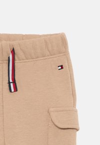 Pantalones de chándal de algodón beige con cintura elástica, que presentan un cordón a rayas y un bolsillo lateral con detalle de costura. Logo de marca pequeño presente.