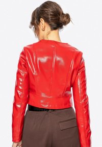 Veste en simili cuir rouge à coupe courte avec une finition brillante, présentant des coutures structurées et des manches longues, associée à un pantalon marron taille haute.
