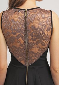 Robe noire avec un dos en dentelle marron et une fermeture éclair. La dentelle présente un motif floral, complété par une jupe noire unie.