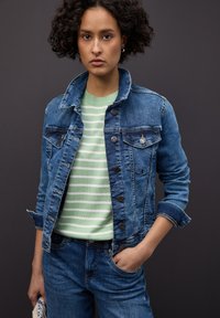 Veste en jean délavée clair, avec deux poches poitrine et des boutons argentés. Portée sur un pull vert rayé. Modèle en jean bleu.