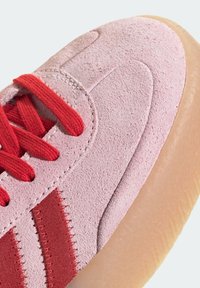 Růžová semišová teniska s texturovaným svrškem, červenými tkaničkami a pruhy. Poloprůhledná gumová podrážka. Detailní záběr ukazuje detaily šití a materiál.