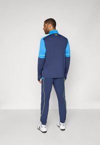 Veste de sport bleu marine avec des manches bleu clair et des accents verts, assortie à un pantalon bleu marine, tous deux en matériau lisse et extensible.