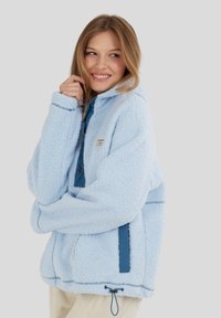 Jonge vrouw die glimlacht en een lichtblauwe fleece jas met marineblauwe accenten draagt, met haar handen dicht bij haar gezicht, tegen een effen lichtgrijze achtergrond.