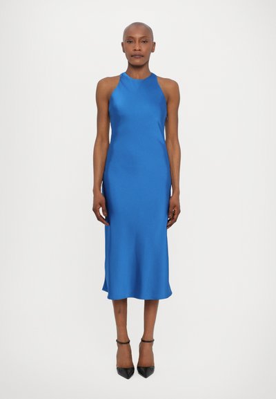 Robe bleue en satin à encolure halter avec une silhouette droite, ourlet au genou et fentes sur les côtés, associée à des escarpins noirs à bout pointu.
