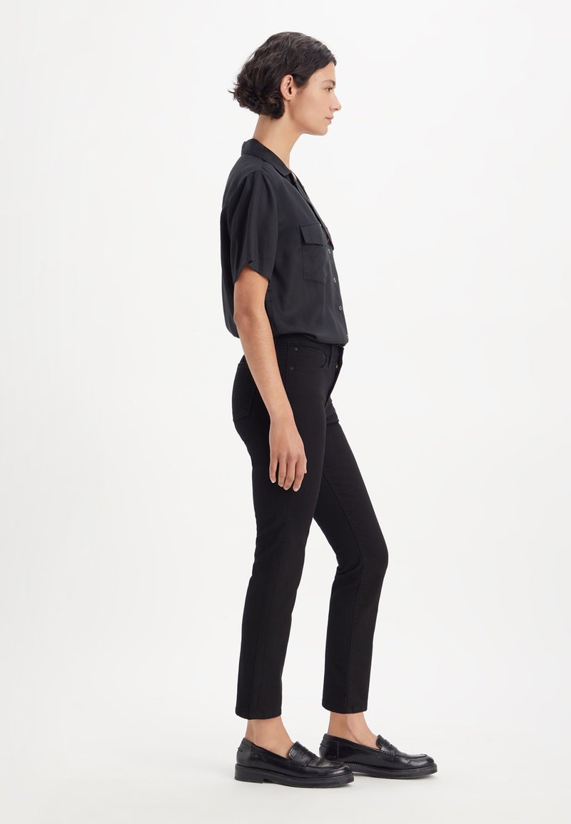 Chemise noire à manches courtes avec deux poches sur la poitrine, associée à un jean noir slim et des mocassins noirs, présentée en profil latéral.