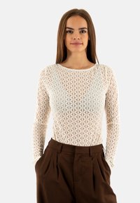 Top a maniche lunghe in rete bianca con un motivo floreale, con scollatura rotonda, indossato con pantaloni marroni a vita alta. Texture liscia.