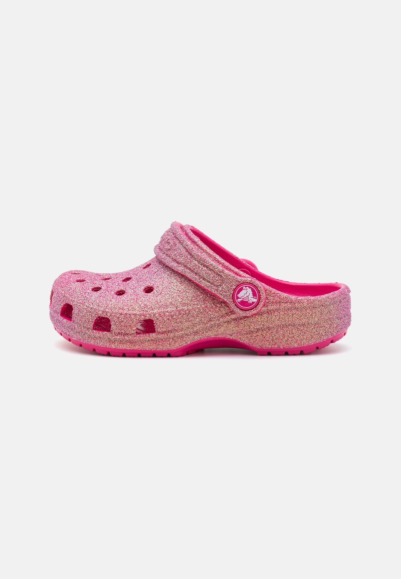 Pink glitter clogs with a rounded design, ventilated holes, and a textured exterior. Features a pink strap and removable logo embellishment.
