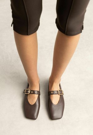 Benen met cropped bruine broeken en donkerbruine Mary Jane schoenen met vierkante neus en gouden gespsluitingen op een lichte vloer.