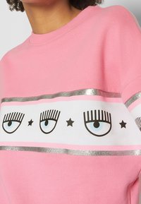 Sudadera rosa con una banda blanca que presenta gráficos de ojos y estrellas negras, destacada por una franja plateada a lo largo del pecho.