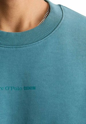 Nahaufnahme von Hals und Brust mit einem blauen gerippten Rundhalsausschnitt-Pullover, der einen schwachen "Marc O'Polo DENIM"-Text und eine silberne Kettenhalskette trägt.