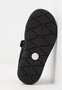 Czarne obuwie z teksturowaną gumową podeszwą o geometrycznym wzorze i okrągłym logo na środku. Szczegół z paskiem na rzep z boku.