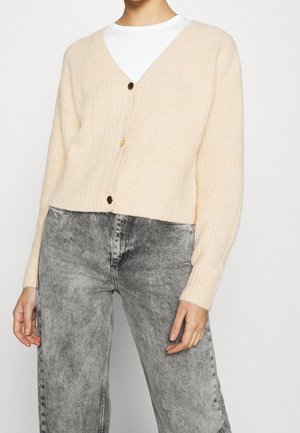 Beige ribbed button-up cardigan over white top paired with high-waisted acid-wash gray jeans on standing person.