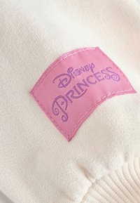 Krémová tkanina s růžovým obdélníkovým dílem, na kterém je vyšitý nápis "Disney PRINCESS" fialkovou nití. Hladký povrch s prošívanými akcenty.