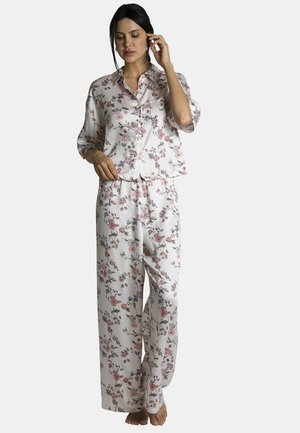 Femme portant un pyjama blanc à motifs floraux avec manches longues et pantalons évasés, debout pieds nus contre un fond uni.