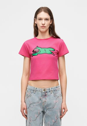 Ženska, oblečena v svetlo roza kratko majico z zelenim risanim živalskim motivom in svetlo modre kavbojke z rožastimi roza vezeninami, stoji na belem ozadju.