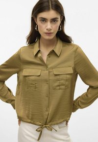 Camicia verde oliva con bottoni, dalla texture leggermente lucida; presenta due tasche sul petto e un nodo all'orlo, contornata da un colletto arrotondato.