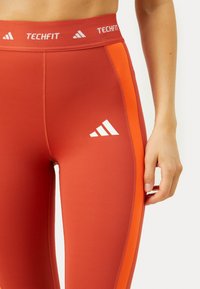 Punased legginsid sobiva disainiga, millel on oranžid külje triibud ja valge Adidas logo. Valmistatud siledast, venivast materjalist.