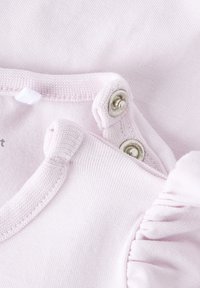 Prenda infantil de color rosa claro con broches de presión plateados, detalles fruncidos y textura suave de algodón. Presenta un escote redondeado y costuras decorativas.