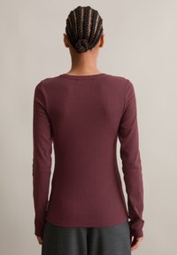 Langermet topp i burgunder ribbet stoff med rund hals, tettsittende stil og myk tekstur. Lapp synlig ved den nedre sømmen.