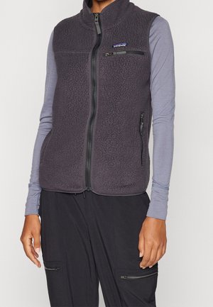 Personne portant un gilet polaire gris foncé sur une chemise gris clair à manches longues et un pantalon noir avec poches zippées, debout devant un fond uni.