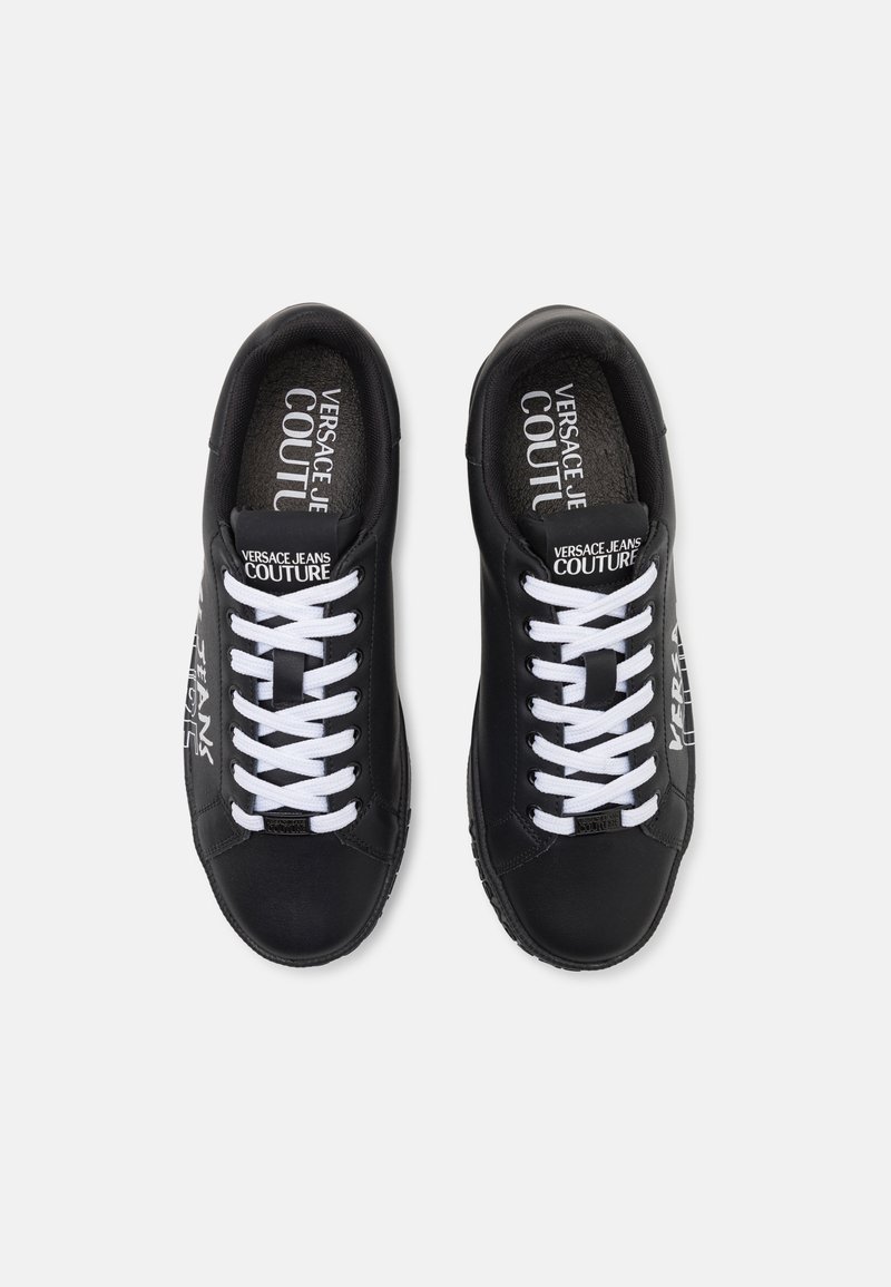 Zapatillas de cuero negro con cordones blancos, que presentan la marca en la lengüeta y el costado, y una suela de goma texturizada. Diseño plano, punta redonda.