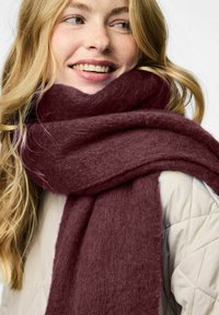 Burgundy tørklæde lavet af blødt, tekstureret materiale, viklet om halsen, med et tykt, hyggeligt design mod en let quiltet jakke.