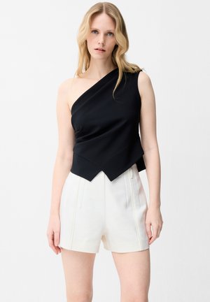 Femme blonde portant un top noir à une épaule avec un ourlet asymétrique et un short plissé blanc taille haute, debout sur un fond blanc.