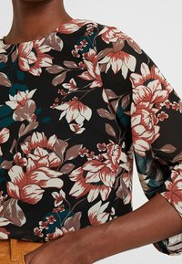 Kwiatowa bluzka na czarnym tle, z dużymi czerwonymi, beżowymi i turkusowymi kwiatami. Miękka tkanina, okrągły dekolt i rękawy o długości trzech czwartek.