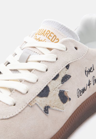 Gros plan sur une basket en daim beige clair avec des lacets blancs, un détail étoile imprimé animal, une semelle marron et un texte de marque orange sur la languette.
