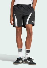 Shorts de sport noirs avec des rayures blanches sur les côtés, taille élastique et poches. Associés à des baskets noires et des chaussettes blanches rayées.